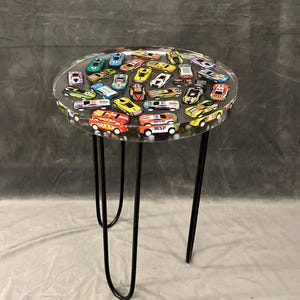 May include: A round clear table with a black metal base. The table top is filled with colorful toy cars embedded in clear resin.