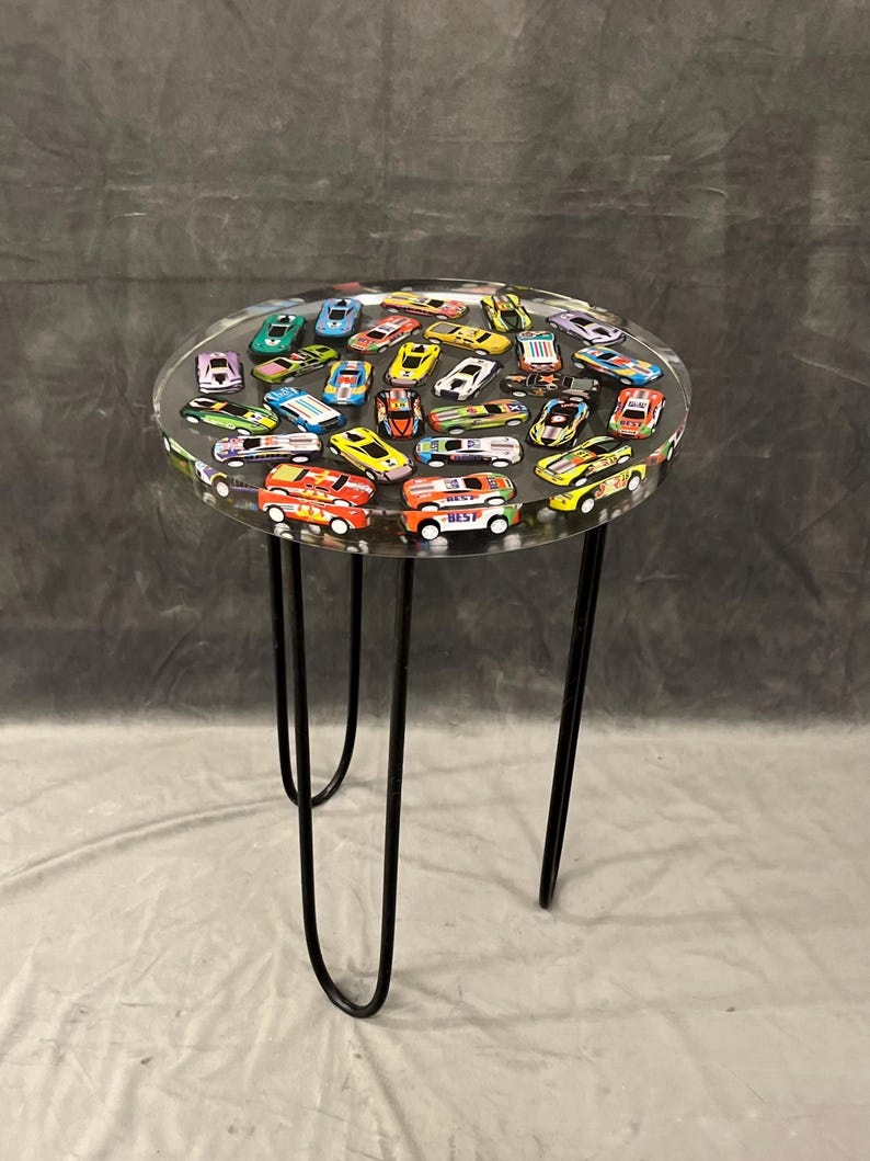 May include: A round clear table with a black metal base. The table top is filled with colorful toy cars embedded in clear resin.