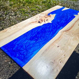 May include: A wooden table top with a blue epoxy resin river running through the center. The wood is a light brown color and the resin is a deep blue with swirls and patterns.