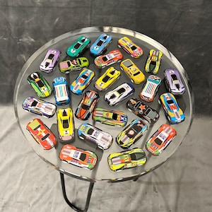 Puede incluir: Una colección de coches de carreras de juguete coloridos con varios números y diseños se muestran en una mesa de vidrio transparente con una base de metal negro.