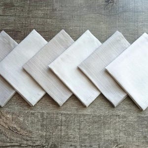 May include: Six square, pale-coloured cloth napkins arranged diagonally on a wooden surface. The napkins vary in shade from light grey to white, with a subtle texture visible. The napkins are likely for dining.