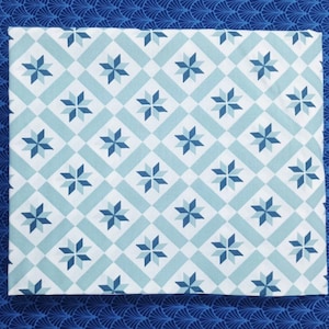 Op de afbeelding: Een vierkante stof met een herhalend patroon van achtpuntige sterren in blauw- en wittinten. De sterren zijn geplaatst in een ruit- en vierkantraster. De achtergrondstof is effen blauw met een waaierachtig ontwerp.