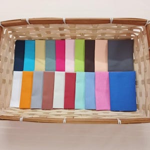 Könnte beinhalten: Ein rechteckiger Weidenkorb gefüllt mit einer Auswahl an farbigen Stoffmustern. Die Muster sind in Reihen angeordnet und zeigen eine Vielzahl von Farben, darunter Blau, Rosa, Grün und neutrale Töne. Der Korb besteht aus hellbraunem Weidengeflecht.