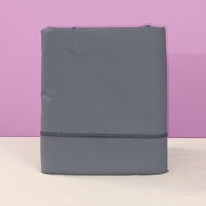 Könnte beinhalten: Ein gefaltetes, einfarbiges graues Bettlaken-Set. Die Bettlaken sind mittelgrau mit einem dunkelgrauen Nahtdetail am unteren Rand. Der Hintergrund ist hellrosa und weiß.