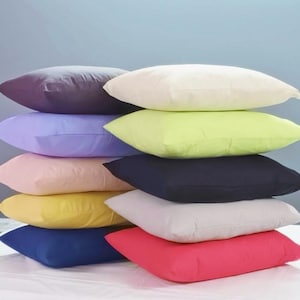 May include: A stack of colorful pillows in various shades, including purple, cream, green, navy, and red. The pillows are rectangular and appear to be made of a soft fabric. They are stacked on top of each other.