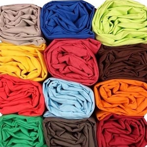 Puede incluir: Una cuadrícula de tela enrollada en varios colores, incluyendo beige, azul, verde, amarillo, rojo, marrón, azul claro, naranja y burdeos. La tela parece ser un material suave, posiblemente algodón o lino.