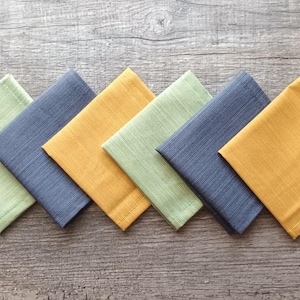 Puede incluir: Seis servilletas cuadradas de tela en fila, cada una de un color diferente. Los colores son verde claro, azul marino, amarillo dorado, verde claro, azul marino y amarillo dorado. Las servilletas son de tela texturizada.