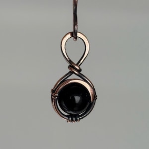 Op de afbeelding: Een koperkleurige hangerketting met een zwarte, ronde edelsteen. De edelsteen is gevat in een draadgewikkelde setting, hangend aan een fijne ketting. Het ontwerp is eenvoudig maar elegant, met een focus op het contrast tussen het metaal en de steen.