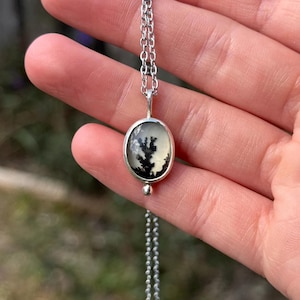 Pu&ograve; includere: Una collana d'argento con un pendente ovale con una pietra di agata dendritica nera e bianca. Il pendente &egrave; appeso a una catena sottile. La collana &egrave; presentata su uno sfondo verde sfocato.