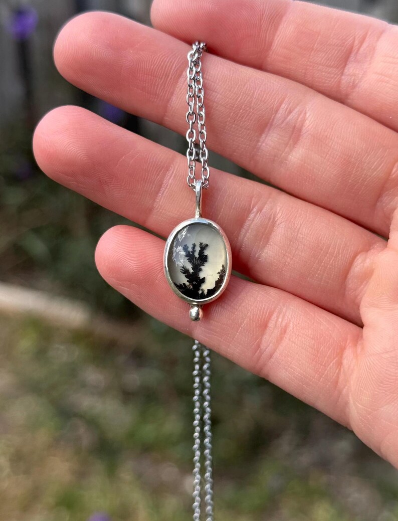 Pu&ograve; includere: Una collana d'argento con un pendente ovale con una pietra di agata dendritica nera e bianca. Il pendente &egrave; appeso a una catena sottile. La collana &egrave; presentata su uno sfondo verde sfocato.