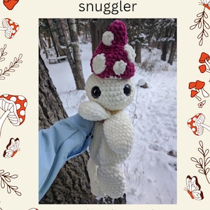 Könnte beinhalten: Ein handgehäkeltes Pilz-Kuscheltier mit weißem Körper und einer magenta-weiß gepunkteten Kappe. Der Text "monty the mushroom snuggle" und "brought to you by cozy critters crochets" sind ebenfalls sichtbar.