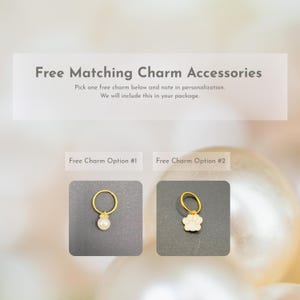 Op de afbeelding: Afbeelding met tekst: "Free Matching Charm Accessories." Er worden twee goudkleurige bedelopties weergegeven. Optie 1 is een gouden ring met een witte parel. Optie 2 is een gouden ring met een witte pootafdrukbedel.