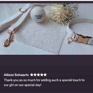 Op de afbeelding: Een witte set voor honden met halsband, riem en tennisbal. De halsband en riem zijn gemaakt van zacht, wit materiaal met goudkleurige hardware. De tennisbal heeft de tekst "with love, Cabela". Een witte roos en decoratieve stof zijn ook aanwezig.