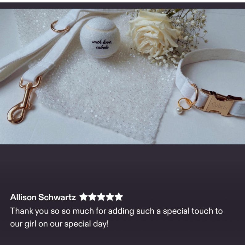 Op de afbeelding: Een witte set voor honden met halsband, riem en tennisbal. De halsband en riem zijn gemaakt van zacht, wit materiaal met goudkleurige hardware. De tennisbal heeft de tekst "with love, Cabela". Een witte roos en decoratieve stof zijn ook aanwezig.