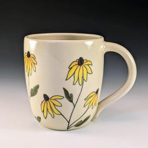 Puede incluir: Una taza de cerámica blanca con un diseño floral. La taza presenta flores amarillas con centros negros y hojas verdes.