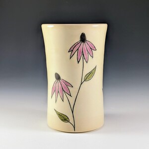 Puede incluir: Un jarrón de cerámica color crema con una forma ligeramente curvada. El jarrón está decorado con dos ilustraciones de flores rosas con centros y tallos negros, y hojas verdes. El diseño está pintado a mano, lo que le da un aspecto artístico único.