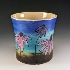 Puede incluir: Una taza de cerámica con un esmalte azul, turquesa y marrón. La taza está decorada con tres flores rosas y moradas con contornos negros.