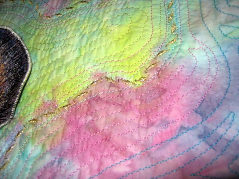 May include: A close-up of a colorful fabric with a quilted texture. The fabric is a blend of pink, blue, green, and yellow hues. The fabric is decorated with gold thread embroidery in a swirling pattern.