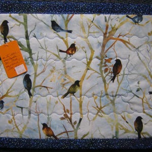 May include: Quilted placemat featuring a watercolor design of birds perched on tree branches. The placemat has a blue border and a tag with handwritten text. The birds are various colors, including brown, blue, and green.