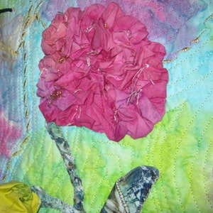 May include: A pink fabric flower with a green and blue background. The flower is made of many layers of fabric and has a ruffled edge. The background is a watercolor wash of green and blue.