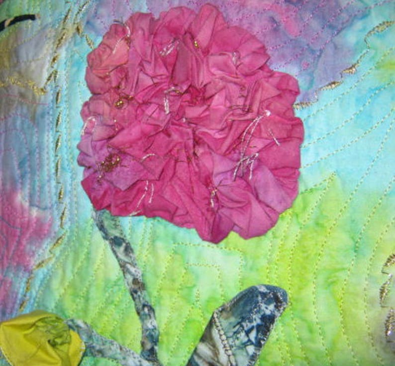 May include: A pink fabric flower with a green and blue background. The flower is made of many layers of fabric and has a ruffled edge. The background is a watercolor wash of green and blue.