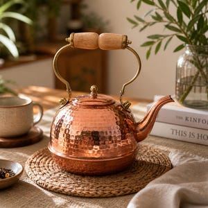 May include: A copper tea kettle with a hammered texture, brass accents, and a wooden handle. The kettle sits on a woven mat, next to a cup and a small bowl. The overall theme is home decor and kitchenware.