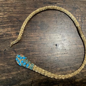 May include: A gold-toned metal snake necklace featuring a textured, scale-like body and a head adorned with small, round, bright blue turquoise-colored stones and two tiny red stones for eyes. The necklace has an open, flexible design.