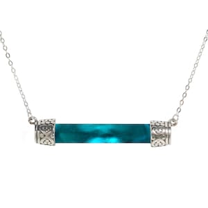 May include: A silver chain necklace with a teal blue glass tube pendant. The pendant has silver caps with intricate designs.