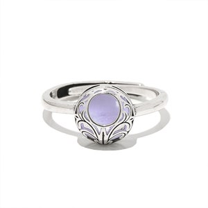 May include: A silver-toned ring with a round, lavender-colored center stone. The stone is surrounded by an ornate, openwork design. The ring is adjustable and sits on a white background.