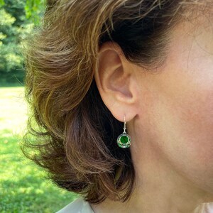 Op de afbeelding: Een paar zilveren oorhangers met een groene edelsteen in het midden van elke oorbel. De oorhangers hebben de vorm van een kleine bol met ingewikkelde zilveren ontwerpen.
