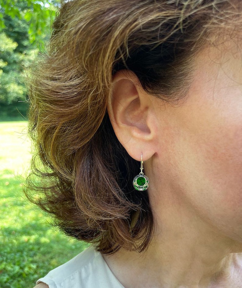 Op de afbeelding: Een paar zilveren oorhangers met een groene edelsteen in het midden van elke oorbel. De oorhangers hebben de vorm van een kleine bol met ingewikkelde zilveren ontwerpen.