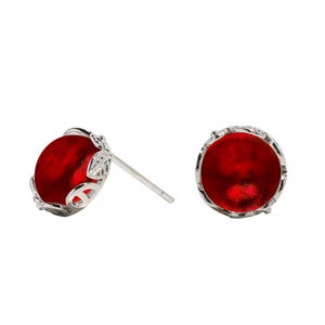 May include: A pair of stud earrings featuring a vibrant, circular red stone set within a polished silver-tone metal frame. The metal setting displays a delicate, openwork leaf-like pattern around the perimeter, with a standard post backing.