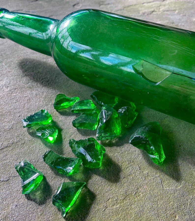 Op de afbeelding: Een gebroken groene glazen fles met scherven glas verspreid eromheen op een grijs oppervlak.