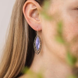 Puede incluir: Un pendiente de filigrana plateado con una piedra ovalada de color lavanda. El pendiente cuelga de un gancho simple. El dise&ntilde;o presenta un patr&oacute;n calado alrededor de la piedra, creando una apariencia ornamentada y delicada. Joyer&iacute;a.