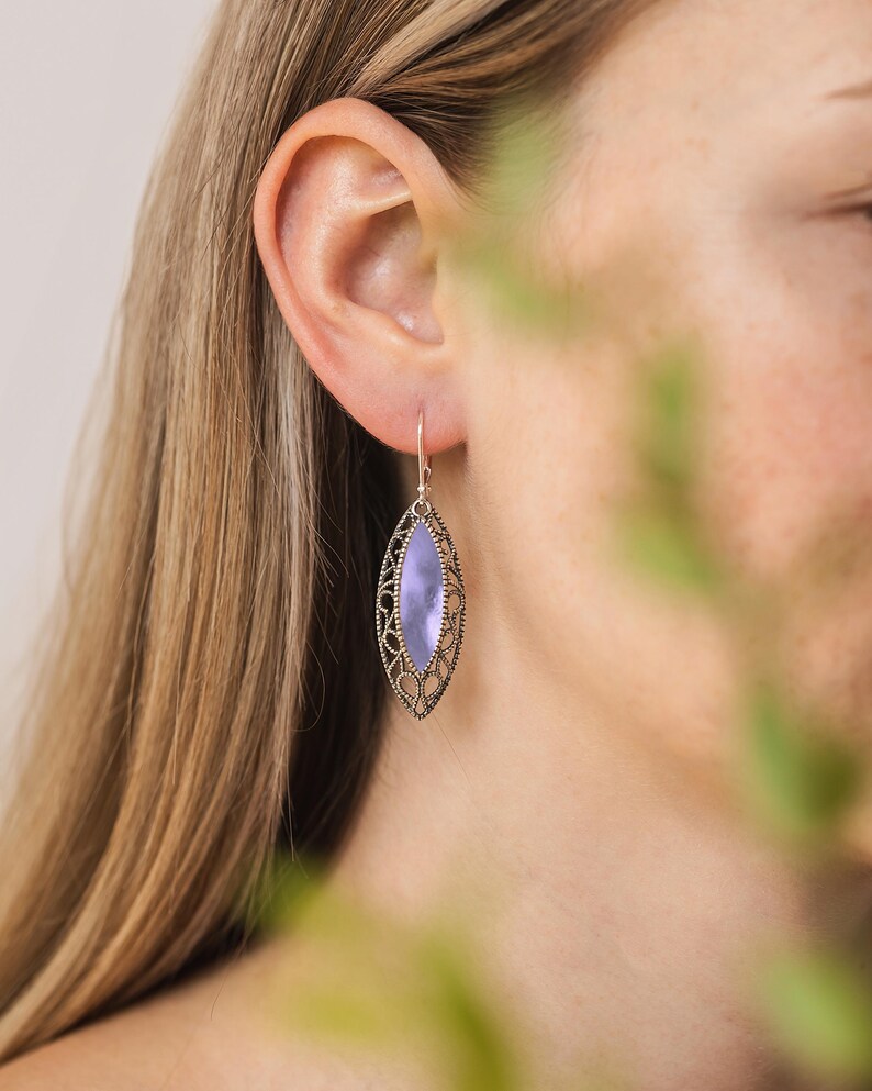 Puede incluir: Un pendiente de filigrana plateado con una piedra ovalada de color lavanda. El pendiente cuelga de un gancho simple. El dise&ntilde;o presenta un patr&oacute;n calado alrededor de la piedra, creando una apariencia ornamentada y delicada. Joyer&iacute;a.
