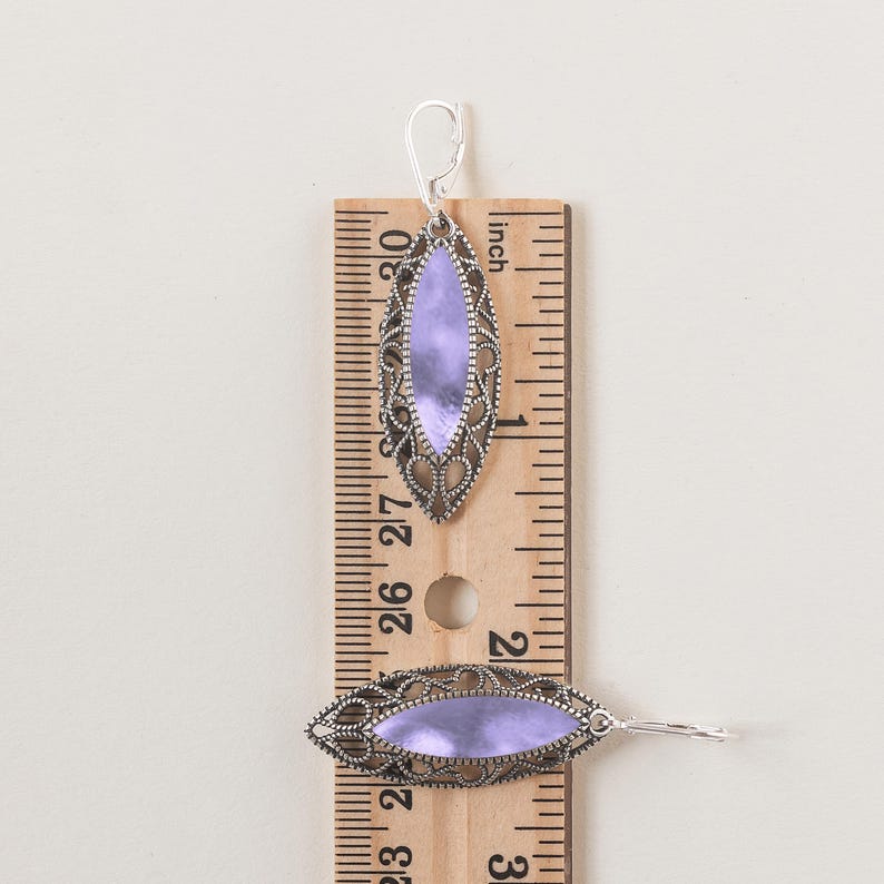 Puede incluir: Pendientes plateados con un centro ovalado de color lavanda. Los pendientes miden aproximadamente 6,35 cm de largo y presentan un dise&ntilde;o calado. Tienen un cierre de palanca. Una regla de madera est&aacute; en el fondo.