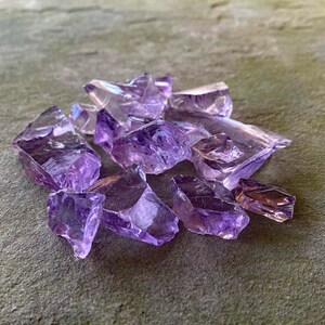 May include: A collection of amethyst crystal chips in various sizes and shapes. The chips are a light purple colour and have a translucent appearance.