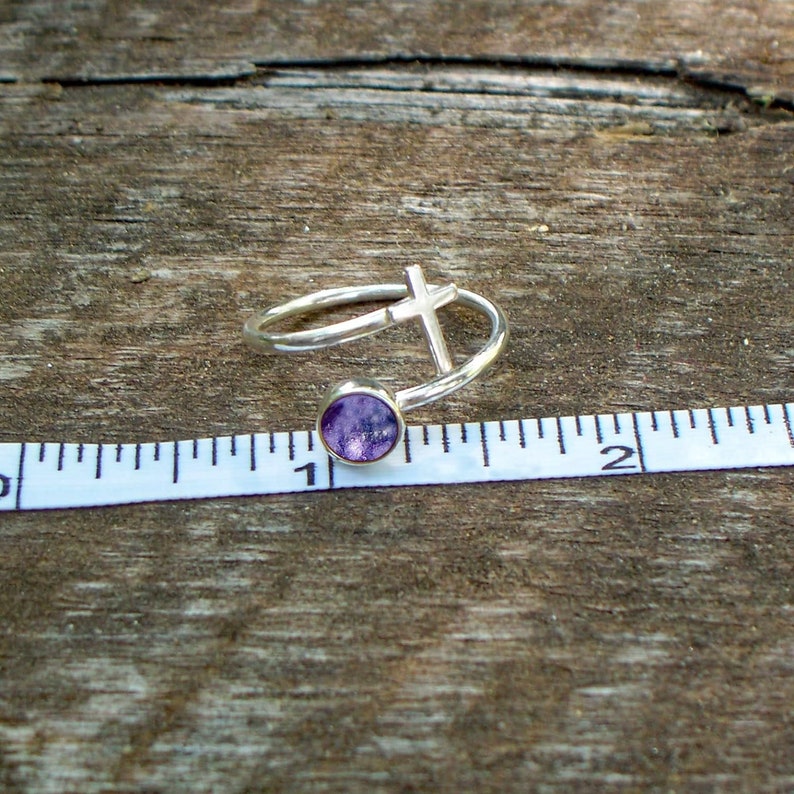 May include: A silver ring with a purple stone and a cross design. The ring is approximately 2.5 cm in diameter.