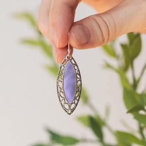Puede incluir: Un solo pendiente plateado con una piedra ovalada de color lavanda. El pendiente tiene un dise&ntilde;o de filigrana abierta alrededor de la piedra. El pendiente est&aacute; sostenido por una mano sobre un fondo borroso verde y blanco.