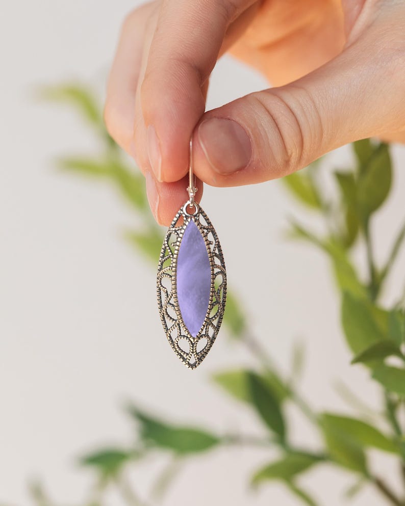 Puede incluir: Un solo pendiente plateado con una piedra ovalada de color lavanda. El pendiente tiene un dise&ntilde;o de filigrana abierta alrededor de la piedra. El pendiente est&aacute; sostenido por una mano sobre un fondo borroso verde y blanco.