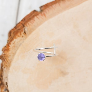 May include: A silver ring with a purple stone and a cross design. The ring is on a wooden surface.