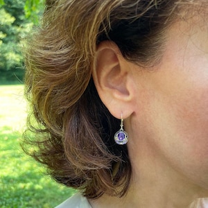 May include: A pair of silver dangle earrings with a purple gemstone in the center of each earring. The earrings are in the shape of a small ball with intricate details.