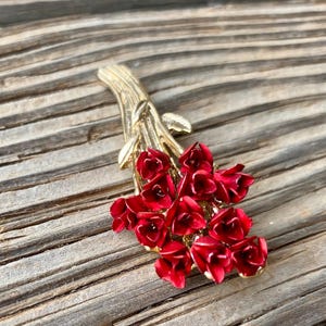 May include: A gold-toned brooch featuring a bouquet of red enamel flowers. The flowers are arranged in a cluster and have a realistic appearance.