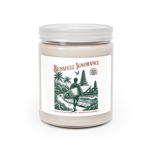 May include: A beige candle jar with a white lid. The label features a green and white illustration of a surfer carrying a surfboard, with palm trees and a temple. The text reads "Blissfull Ignorance" and "It's all fun and games until someone gets murdered."
