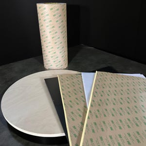May include: A roll of 3M double-sided tape stands upright next to a wooden round surface and several sheets of black and white material. The tape roll is covered in a repeating pattern of the 3M logo.