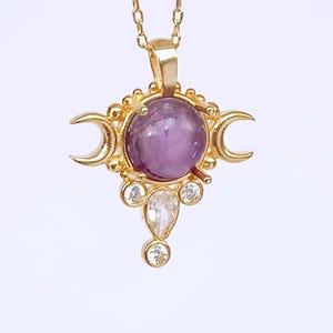 May include: Gold-toned pendant necklace featuring a central purple gemstone, surrounded by crescent moon shapes and smaller clear gemstones. The necklace has a delicate chain.
