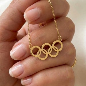 May include: A gold-coloured necklace featuring the Olympic rings design. The necklace is held in a hand, showcasing the delicate chain and the five interconnected rings. The rings are in a classic Olympic arrangement.
