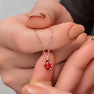 Könnte beinhalten: Eine zarte Halskette mit einem kleinen Marienkäfer-Anhänger. Der Marienkäfer ist rot mit schwarzen Punkten und einem schwarzen Kopf. Die Kette ist roségoldfarben. Die Halskette wird von den Fingern einer Person gehalten.