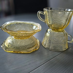 May include: Two vintage yellow glass cups with a floral pattern. The cups are stacked on top of each other. The cup on top has a handle. The cups are sitting on a gray wooden surface.