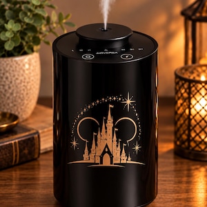 Puede incluir: Un humidificador cilíndrico negro Airversa que emite un chorro de vapor vertical. El frente presenta un gráfico dorado de la silueta de un castillo enmarcado por dos círculos y estrellas. El panel de control superior incluye iconos.